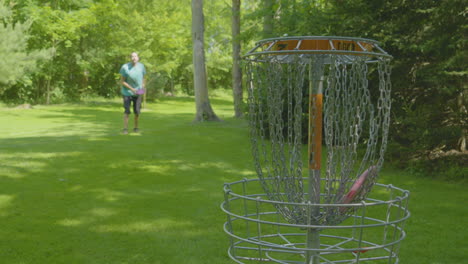 a man celebrates a successful putt as his disc lands in the basket, set in a lush green park, capturing the joy of disc golf