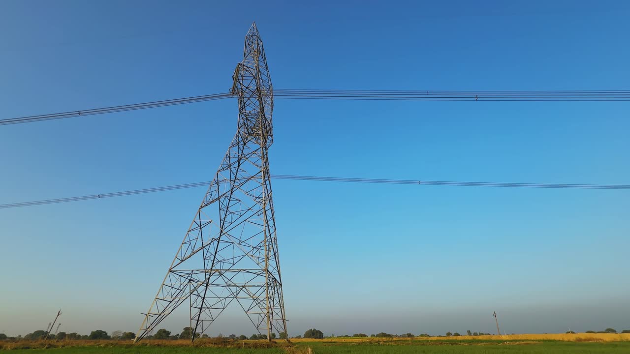 Tilt-up view from green crop field toward tall electricity transmission tower,steel lattice structure and power lines against clear blue sky,rural energy infrastructure intersecting agriculture
