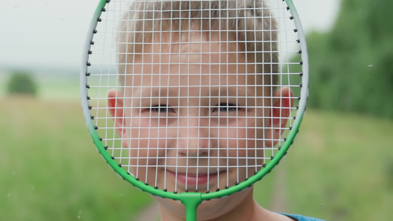 코카서스인 아이가 필드에서 라켓 사이로 빼꼼히 쳐다보는 모습, 초록색 스트링 뒤로 보이는 미소와 역동적인 클로즈업, 여름 미풍과 흐릿한 시골 배경, 솔직한 스포츠 표정을 짓는 어린 운동선수의 분위기