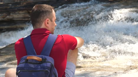 solo viajero masculino con mochila se sienta cerca del río de montaña burbujeante