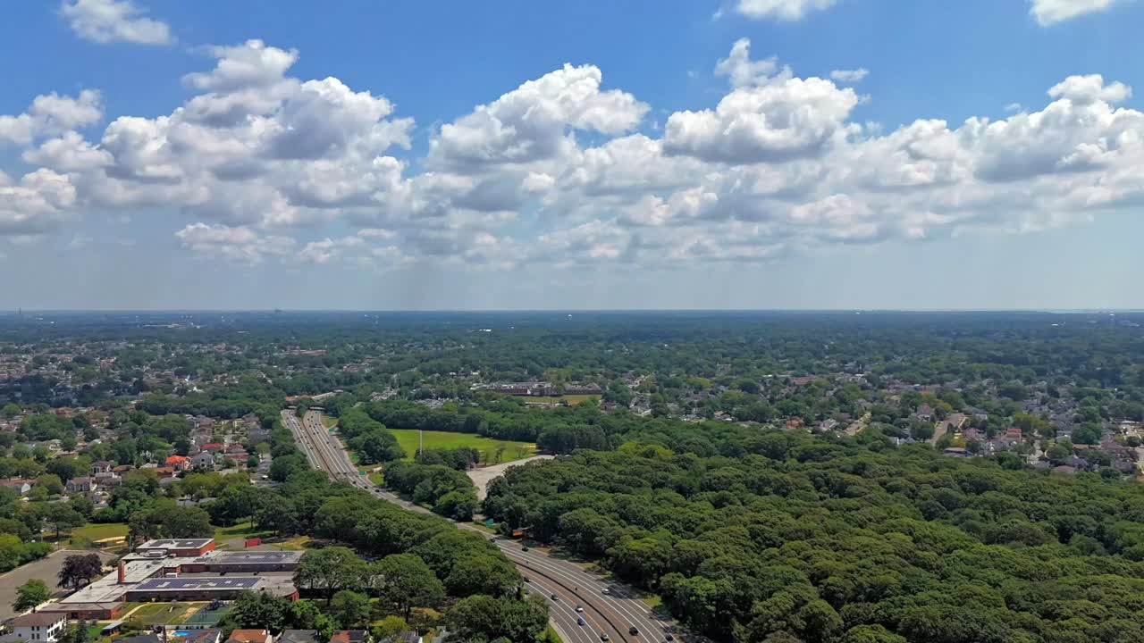 An aerial time lapse over Southern State Parkway on Long Island during a sunny day. Flying over green trees in the suburban neighborhood. The camera dolly out and tilt down over the landscape