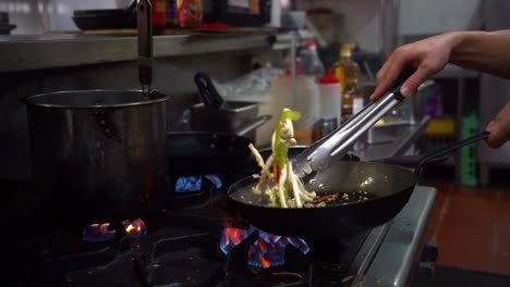 professional chef flipping the ingredients in the frying pan and mixing with a tongs, cooking delicious veggie and mix mushrooms for stir frying spaghetti aglio e olio, slow motion shot