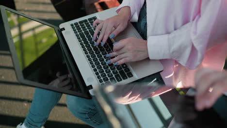 vista de primer plano de una mujer escribiendo en una computadora portátil al aire libre, con uñas pulidas visibles, al lado de una persona que usa una tableta, con sombras de proyección de luz solar