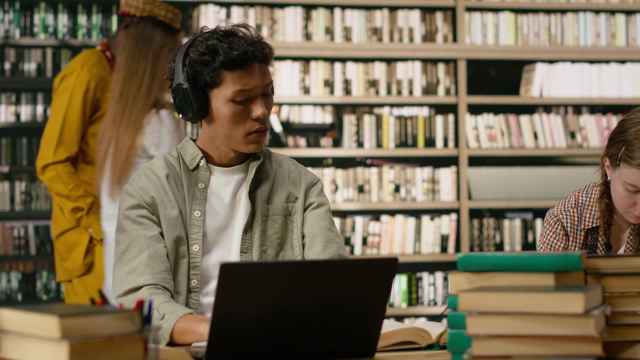 Studente asiatico che usa le cuffie e prende appunti con un portatile e un libro aperto.
