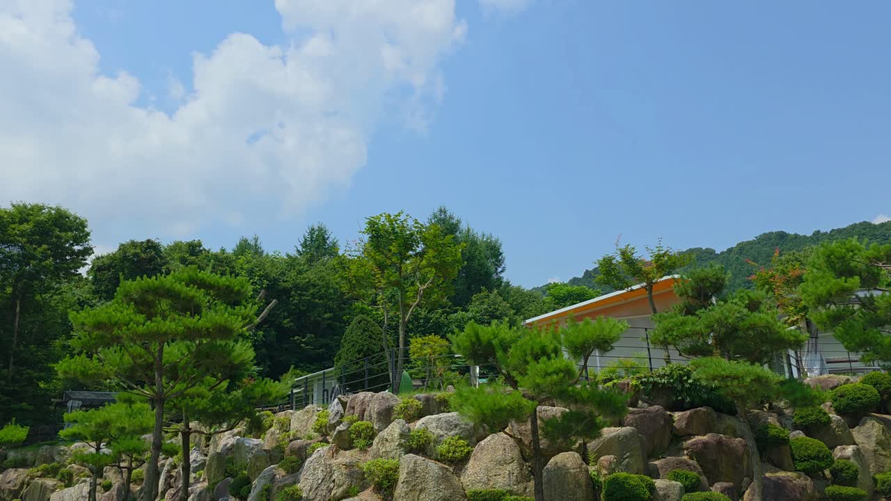 A timelapse captures white fluffy clouds drifting across a blue sky above modern glamping buildings nestled in lush greenery at a South Korean resort