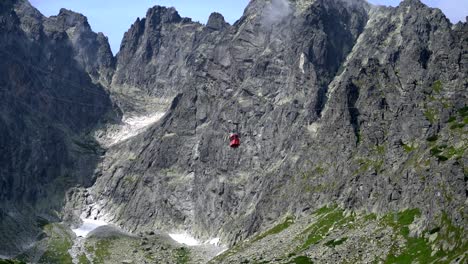 a tiny red gondola slowly descends in front of rugged jagged cliffs, the high tatra mountains