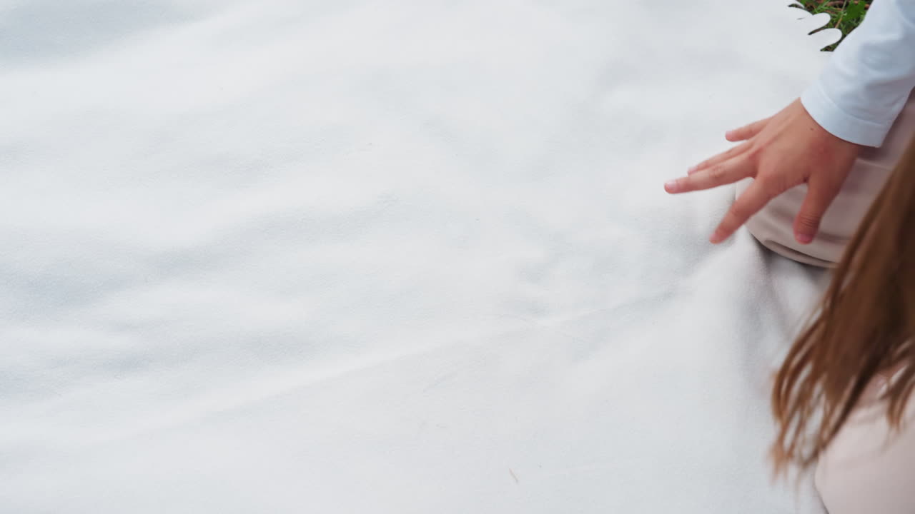 어린 소녀가 피크닉 준비를 하는 동안 초록색 잔디 위로 흰 천을 부드럽게 펴는 모습이에요. 은은한 움직임, 차분한 집중, 따뜻하고 평화로운 분위기를 연출하는 자연광을 보여줍니다.