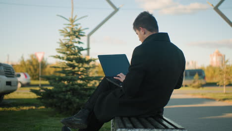 freelancer seated outdoors working on laptop with leg crossed in peaceful urban setting, background includes moving car, parked car, nearby structures, and greenery