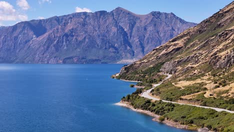 4k drone video of a biker driving around the winding roads at Lake Hawea in Wanaka, New Zealand. The lake is surrounded by towering mountains and large hills filled with trees and grassy landscapes