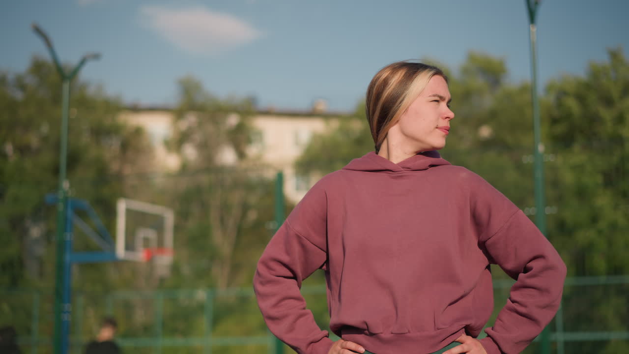 dama con capucha marrón con la mano en la cintura, haciendo ejercicio mientras gira la cabeza de izquierda a derecha, la luz del sol brilla sobre ella con un aro de baloncesto en el fondo