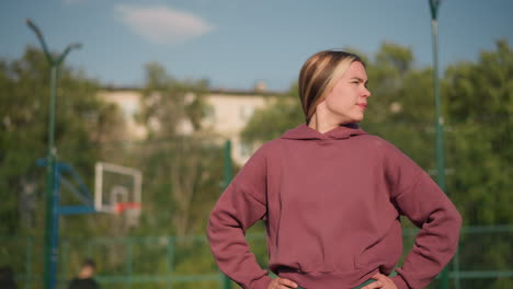 lady in maroon hoodie with hand on waist, working out while turning head from left to right, sunlight shines off her with basketball hoop in background