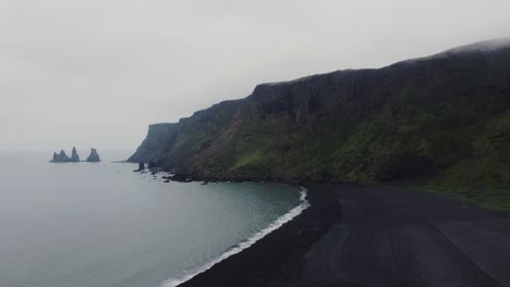 Luftdrohnenaufnahme-Von-Wunderschönen-Blauen-Wellen,-Die-An-Einem-Schwarzen-Sandstrand-In-Island-Zusammenbrechen,-Und-Riesigen-Felsformationen-In-Der-Ferne