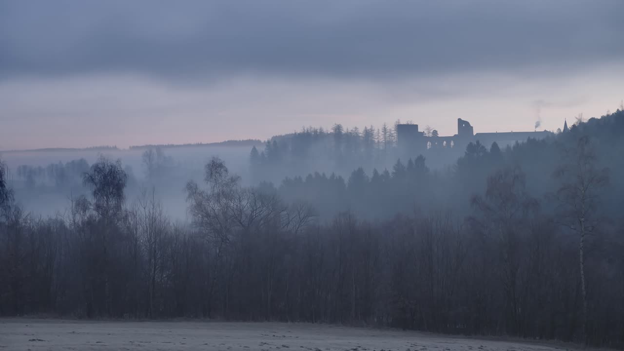 paisaje de niebla tranquila con árboles desnudos, un campo congelado y la silueta de un castillo medieval en una colina, todo bajo un cielo nublado cambiante en una toma de lapso de tiempo