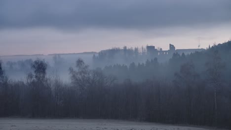 quiet misty landscape with bare trees, a frozen field and the silhouette of a medieval castle on a hill, all under a changing overcast sky in a time-lapse shot