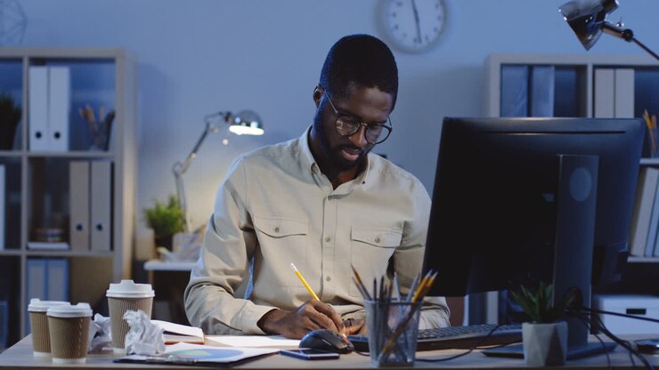 Angry Young Man In Glasses Working With Computer And Writing Something, Then Throws A Sheet Of Paper Away In The Office At Night