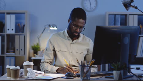 Angry-Young-Man-In-Glasses-Working-With-Computer-And-Writing-Something,-Then-Throws-A-Sheet-Of-Paper-Away-In-The-Office-At-Night