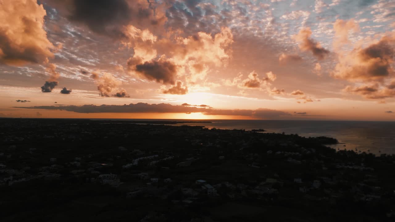Bredt luftpanorama, der fanger den livlige, skyggefulde solnedgang over den mørke, silhuetklædte mauritiske kystlinje og boliglandskab. Konceptet formidler tropiske aftener og fredfyldte rejseøjeblikke.