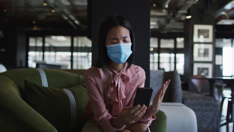 Asiatische-Frau-Mit-Gesichtsmaske-Führt-Einen-Video-Chat-Auf-Dem-Smartphone-Im-Modernen-Büro