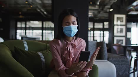 asian woman wearing face mask having a video chat on smartphone at modern office