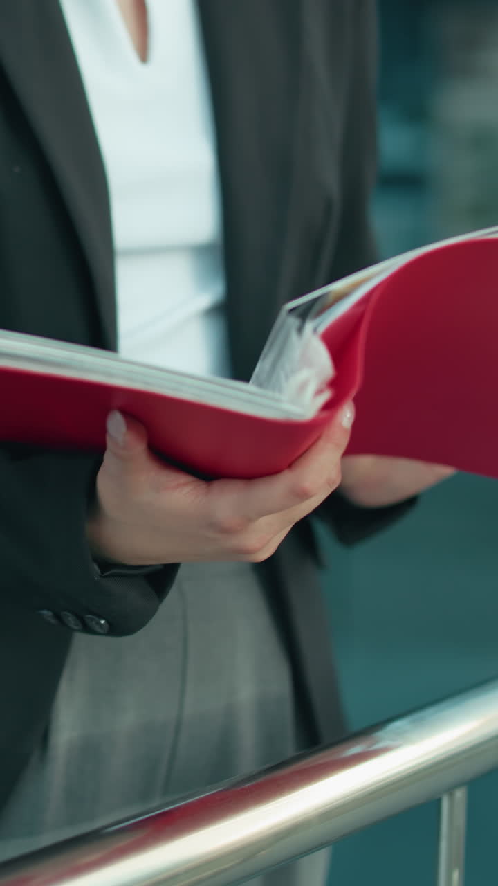 Částečný pohled na dámu v černém saku, která venku zkoumá červenou složku s městskou obytnou budovou v pozadí, s důrazem na ruce a dokument, což odráží profesionální pracovní prostředí.