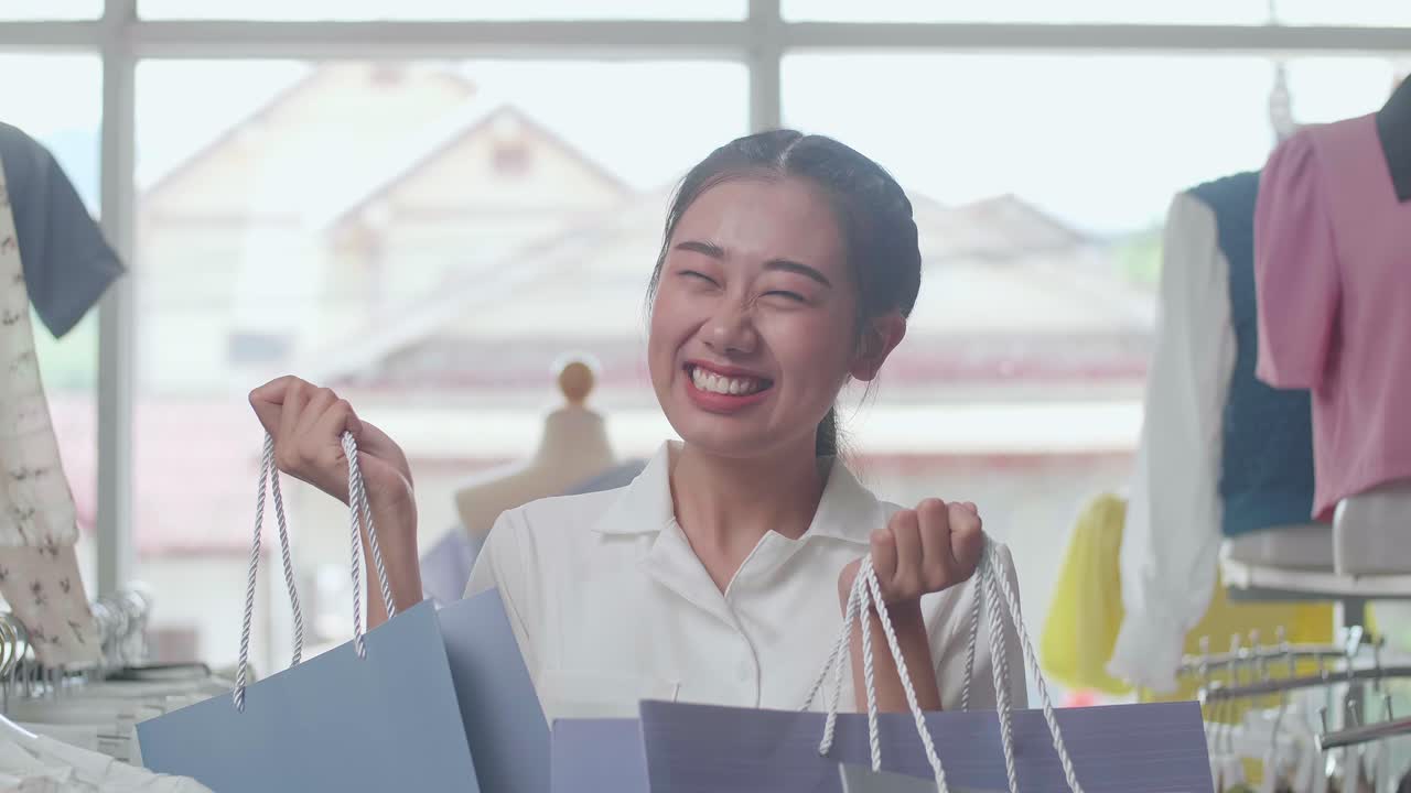 쇼핑 가방을 들고 춤추며 축하하는 즐거운 젊은 아시아 여성은 부티크에서 쇼핑하는 동안 옷을 살 수 있습니다.