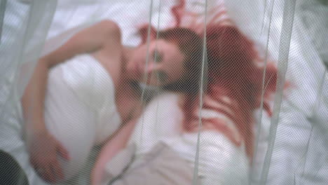 redhead woman lying on white fabric behind transparent veil