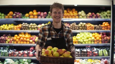 ritratto del giovane bellissimo operaio del negozio caucasico nel grembiule in piedi davanti alla telecamera e sorridendo gioiosamente mentre tiene in mano una scatola di mele al supermercato. sfondo del pannello di frutta e verdura