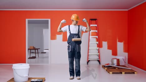 full body of asian man painter wearing safety helmet smiling and flexing his bicep while standing with painting wall in room