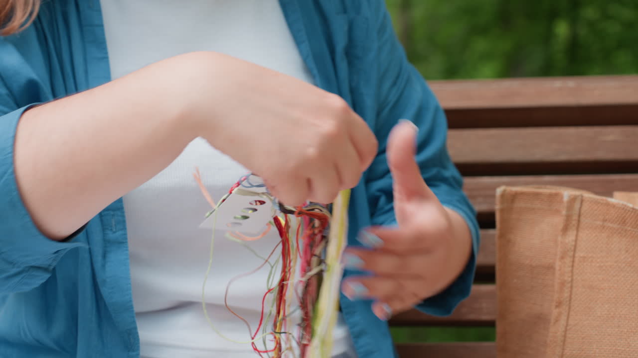 Lady sitting on wooden bench outdoors unwraps colorful embroidery thread bundle, dropping loose threads beside her while preparing for creative crafting project in calm natural daylight atmosphere