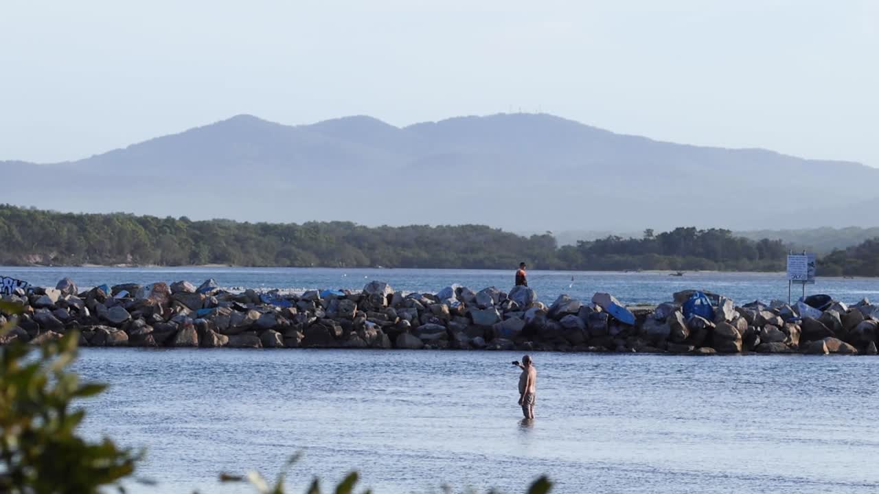 A tranquil scene of a rocky shoreline with distant mountains and a lone figure standing in the water.