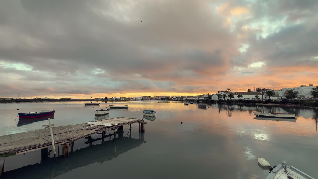 Stunning sunrise illuminating barbate's tranquil harbor waters, reflecting vibrant skies and clouds, while small fishing boats sway near a weathered wooden pier in andalusia, spain