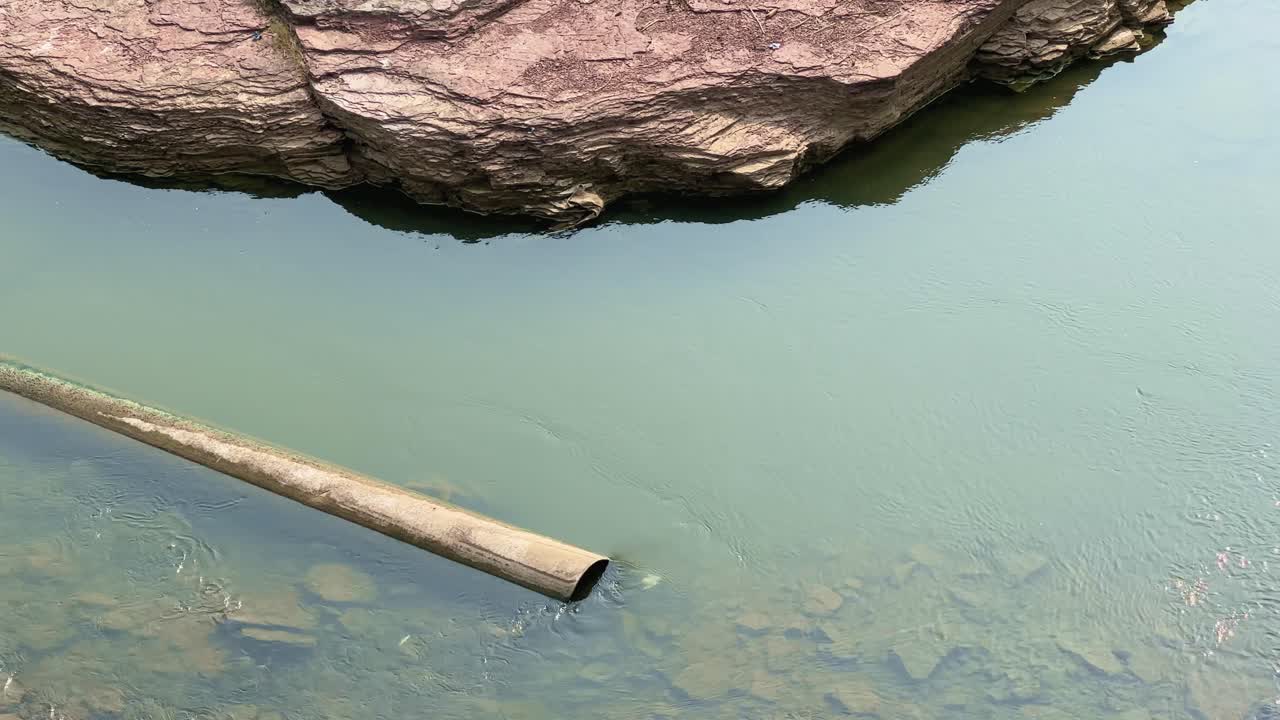 A close-up view of a rusty water pipe extending into a calm green river beside large rocky formations, showing rural water usage and the blend of natural and human-made elements in a quiet environment