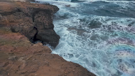 rough waves crash against the rocky cliffs with a cave-like formation visible, creating a dramatic natural scene along the coast