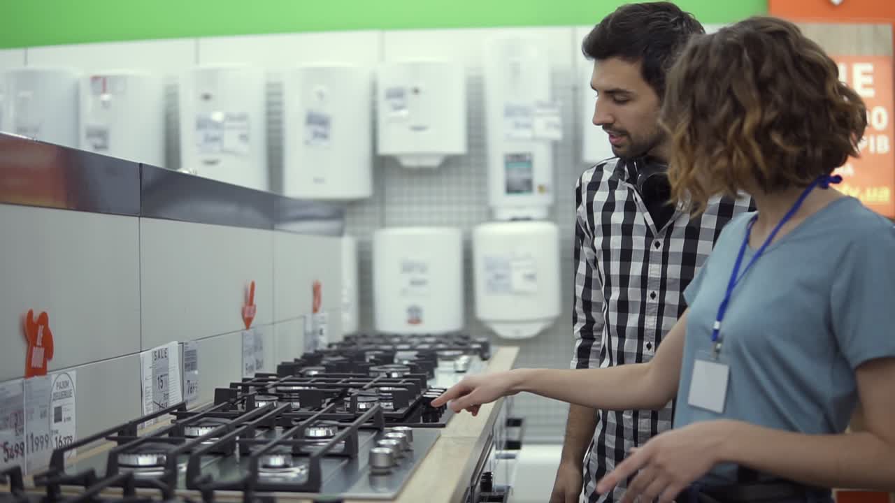 Male shopper in plaid shirt with headphones choosing a new modern oven in store. He is planning to buy furniture and home appliances, checking and advising with female consultant