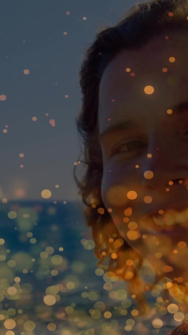 Video vertical: Mujer inclinándose hacia el mar, sonriendo con destellos dorados de bokeh cubriendo su rostro, para contenido de viajes.