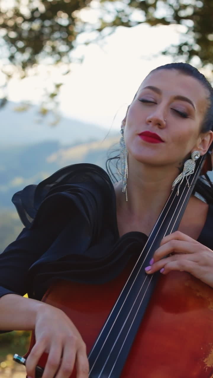 Mid-aged young woman with makeup and nice black dress performs cello music. Lady closed her eyes enjoying music. Nature backdrop in blur. Vertical video