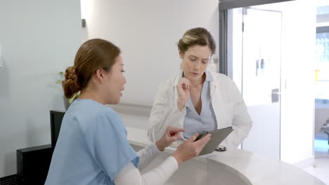 focused diverse female doctors using tablet and discussing over hospital reception desk, slow motion