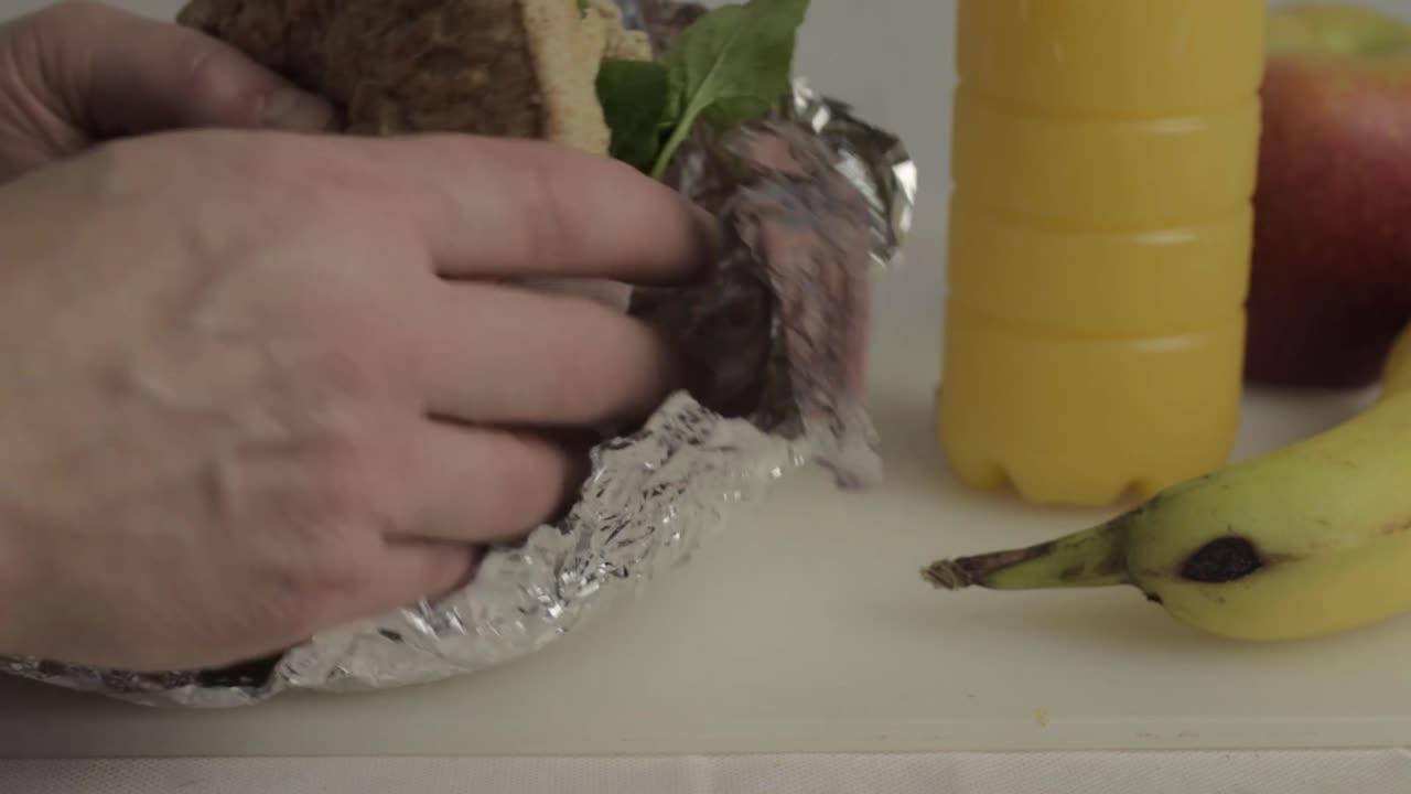 Unpacking a foil wrapped sandwich with orange juice and fruit close up