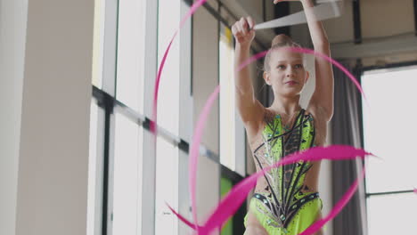 smiling young girl in leotard practising rhythmic gymnastics with a ribbon in a studio