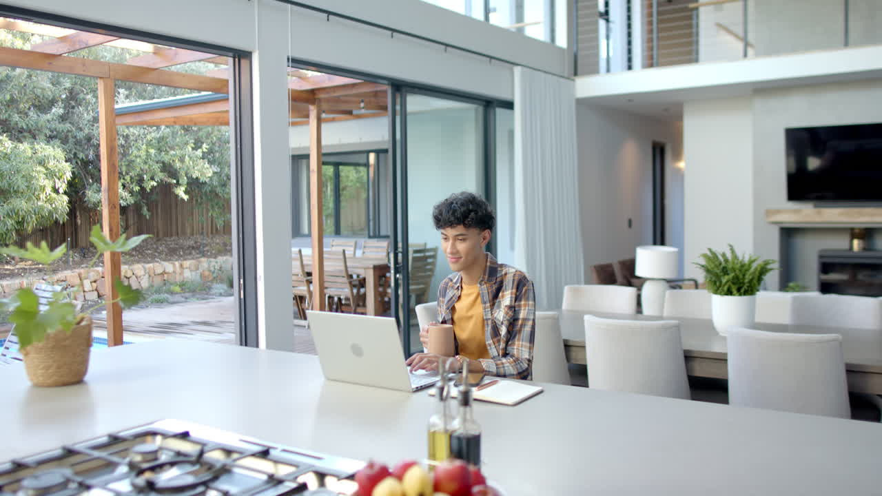 Working from home, teenage boy using laptop and holding coffee mug in kitchen