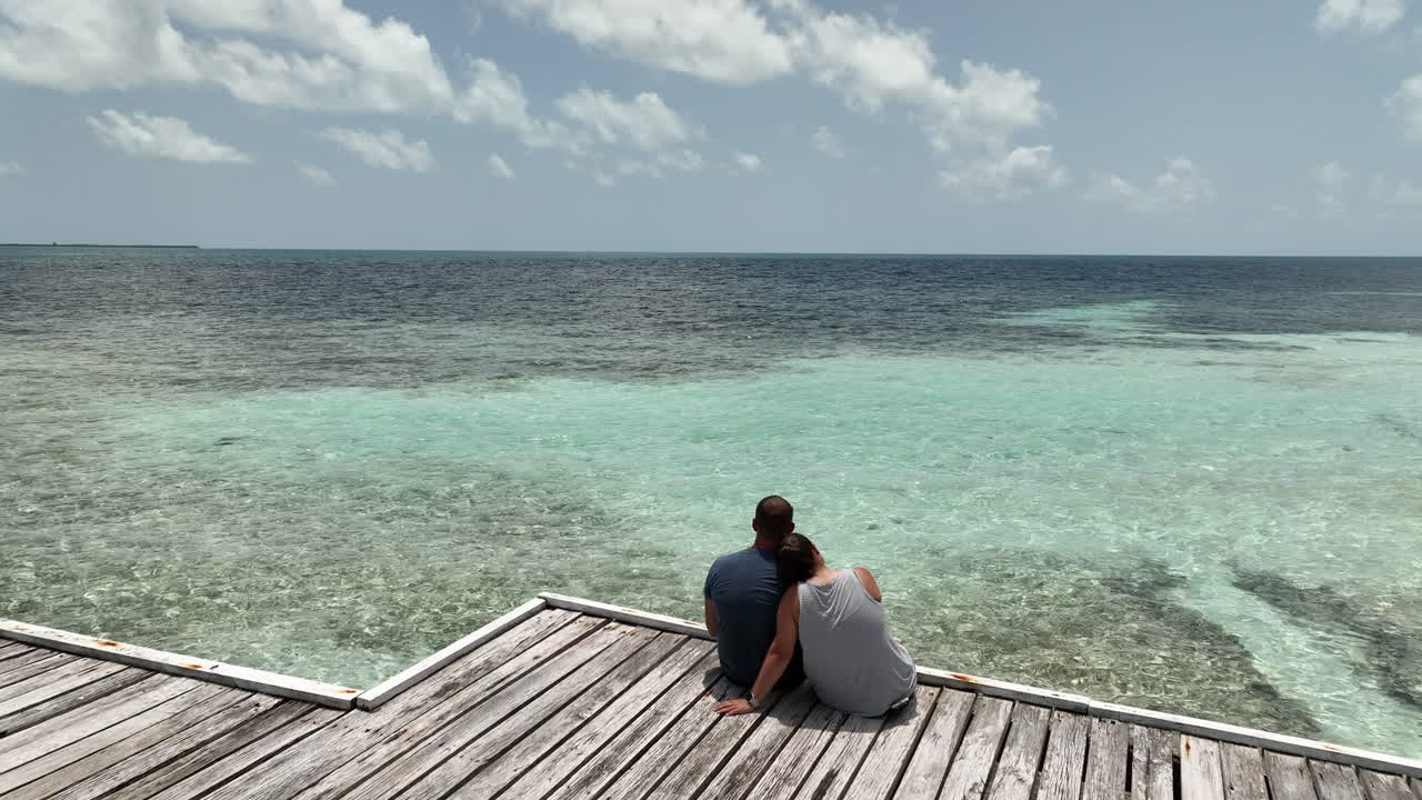 Behind passover aerial view of couple sitting on pier overlooking the Caribbean.