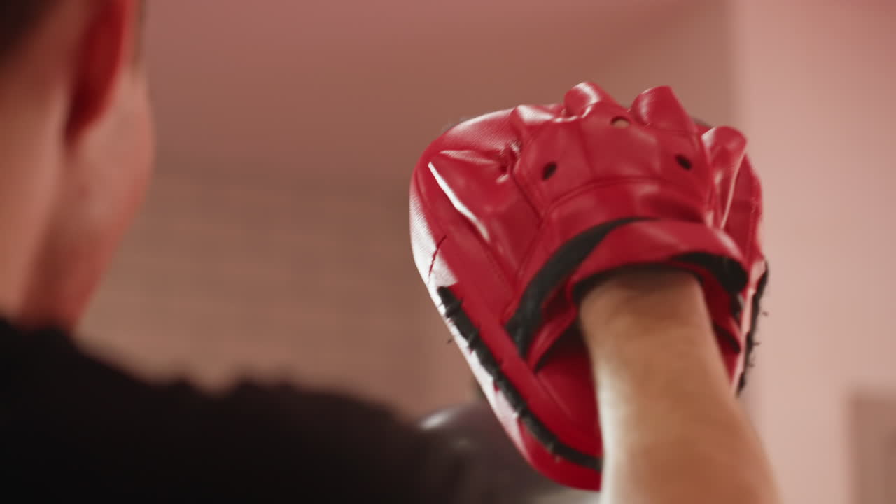 Rear view of boxer trainer holding red boxing pad during sparring practice, showing focus on equipment, hand grip, and preparation for striking training session in gym with intense atmosphere of combat sports