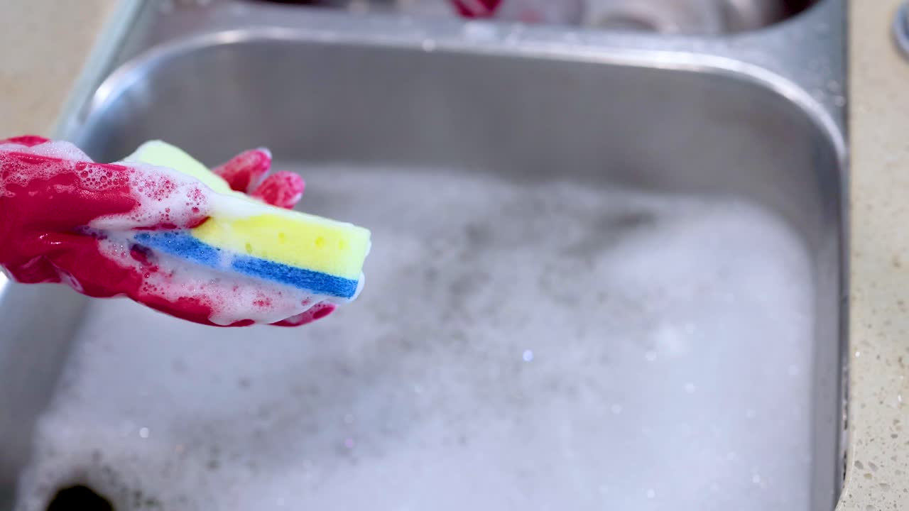 Hands in red gloves clean a glass with a sponge over a soapy kitchen sink