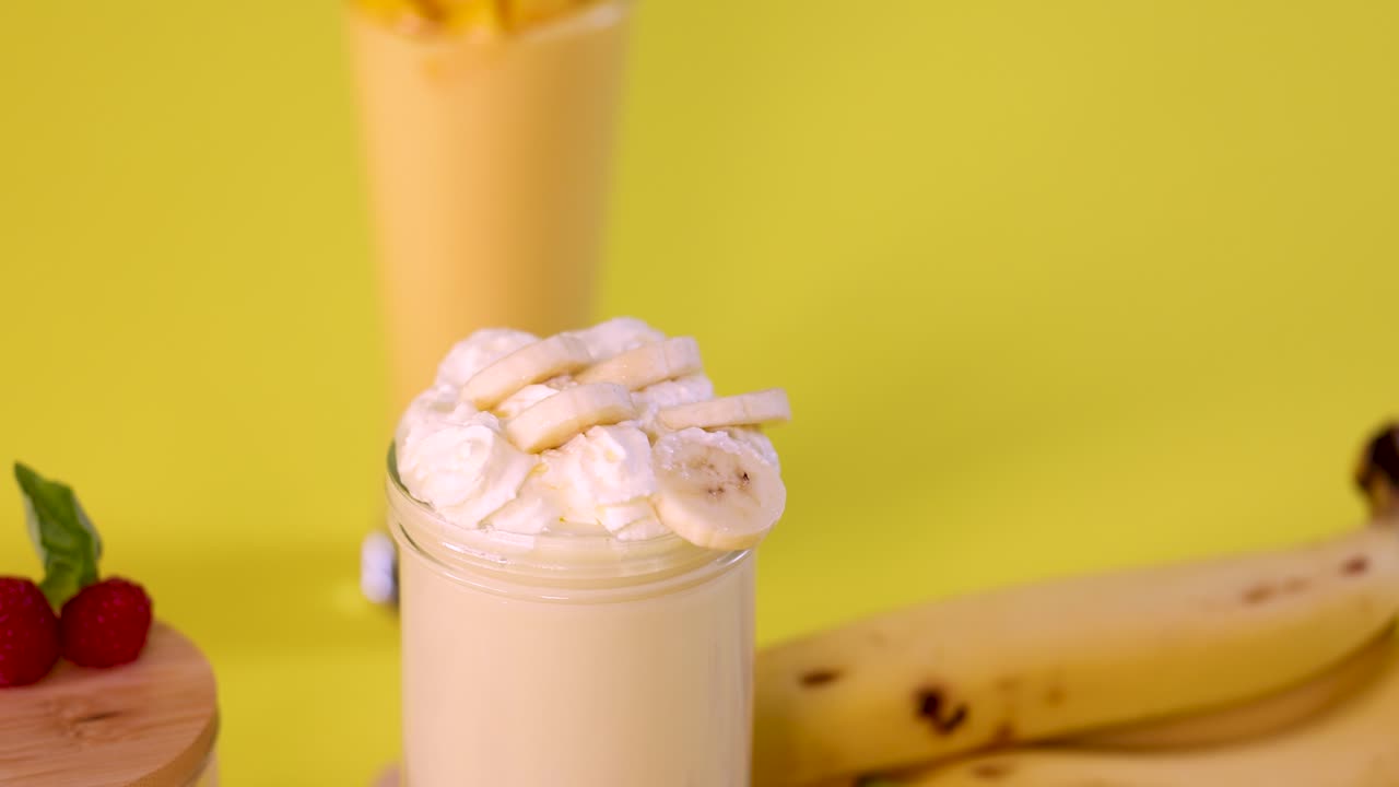 Banana milkshake topped with banana slices, colorful sprinkles falling, bright yellow background, static camera