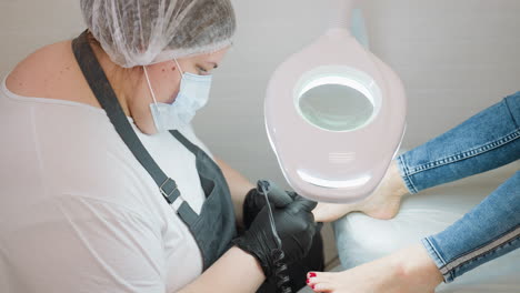 beautician wearing mask and gloves using electric filer to shape toenails of client sitting on treatment chair under bright salon lamp during professional pedicure procedure in clean indoor setting