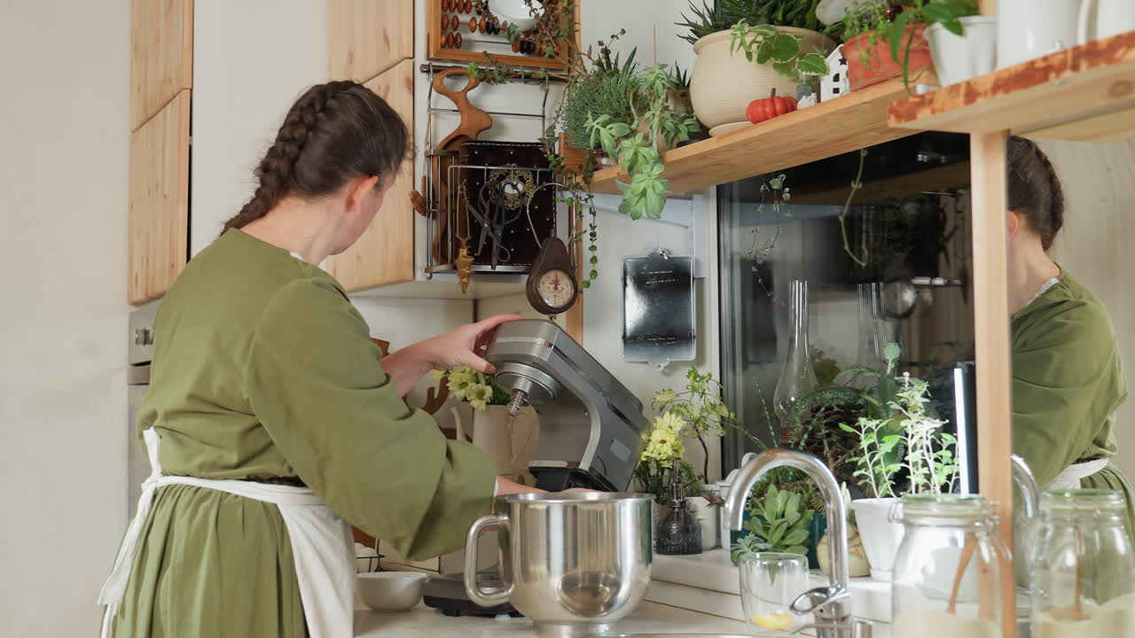 Woman in green gown and white apron opens kitchen blender in cozy modern kitchen surrounded by greenery and utensils, preparing ingredients in stainless bowl reflected in wall mirror