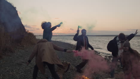 grupo de amigos adolescentes corriendo alrededor de una hoguera sosteniendo bengalas de colores en la orilla del mar 1