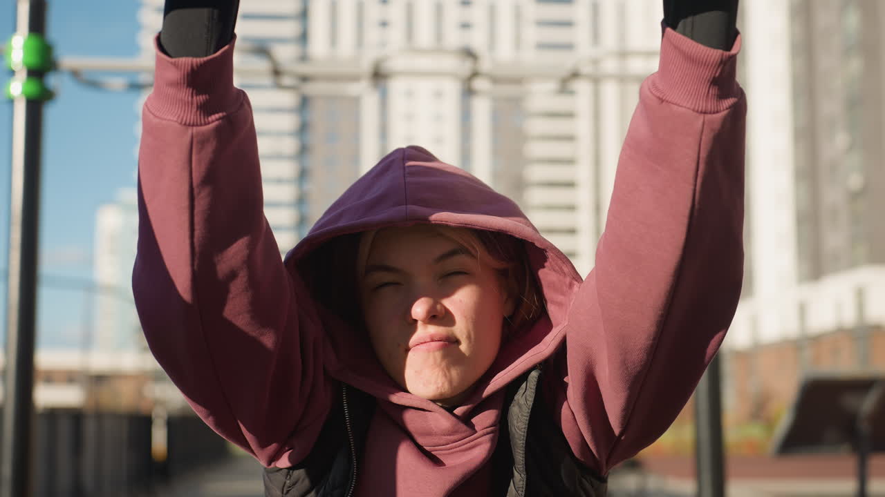 Fitness trainer gripping bar under sunlight, resilience on face, hoodie wearing athlete performing pull exercise on outdoor station, urban backdrop with exercise rails and high rise buildings