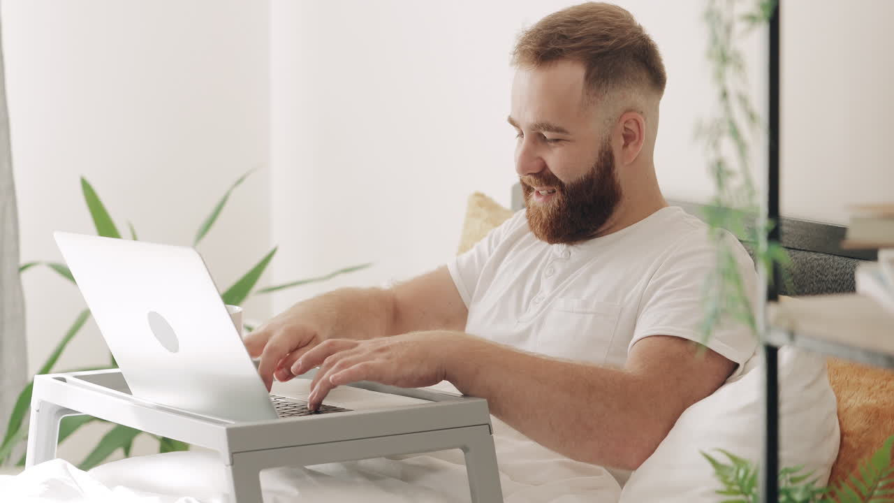 Бородатый мужчина пьет кофе и работает за ноутбуком в постели