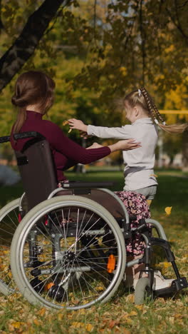 glad mother in wheelchair extends hands to dancing daughter. cheerful preschooler girl scatters yellow leaves spinning and leans hugging mum lovingly